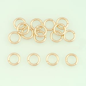 Op de afbeelding: Een verzameling goudkleurige, cirkelvormige sieradssluitingen, gerangschikt op een lichtgroene achtergrond. De sluitingen zijn rond met een scharnierend ontwerp, wat suggereert dat ze voor oorbellen of andere sieraden zijn. De afbeelding toont de sluitingen in een studio.