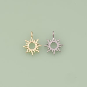 May include: Two sun-shaped pendants. One is gold-colored, the other is silver-colored. Each pendant has a circular center with radiating rays and a small loop at the top for attaching to a chain or other jewelry component. The background is a light green color.