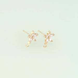May include: A pair of gold-colored stud earrings, each featuring a flower design with white gemstone petals. The earrings have a post and a small, curved loop at the bottom. The background is a soft, light green.