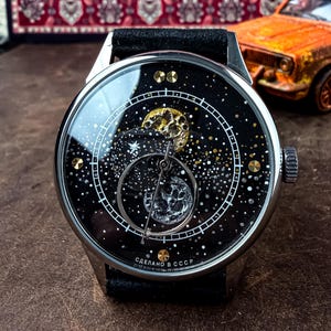 May include: A round wristwatch with a black leather strap and a silver-tone metal case. The dial features a dark space-themed background with gold and silver celestial bodies, a circular sub-dial, and the text "СДЕЛАНО В СССР" at the bottom.