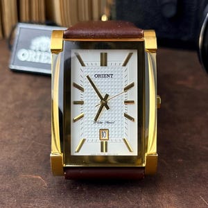 Puede incluir: Reloj Orient rectangular dorado con esfera blanca y correa de cuero marrón. La esfera del reloj presenta agujas y marcadores dorados, con la fecha en la posición de las 3. El reloj es resistente al agua.