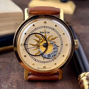 Könnte beinhalten: Goldfarbene Armbanduhr mit braunem Lederarmband. Das Zifferblatt zeigt ein Sonnen- und Monddesign mit dem Wort "RAKETA". Der Text "СДЕЛАНО В СССР" steht unten. Eine Vintage-Uhr.