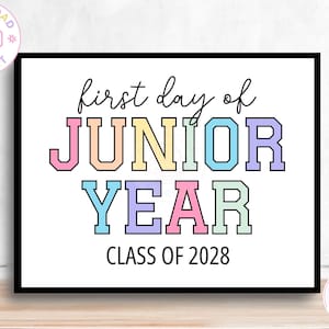 May include: A rectangular sign with a white background and a black border. The text reads first day of JUNIOR YEAR in a mix of cursive and block lettering. The letters are colored in pastel pink, orange, yellow, blue, and purple. CLASS OF 2028 is below.