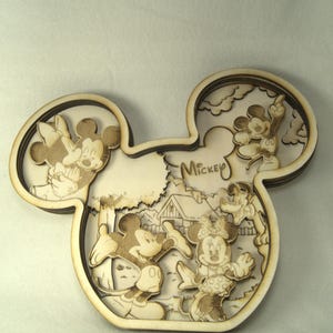Könnte beinhalten: Ein Holz-Ausschnitt von Mickey Mauses Kopf mit einem mehrschichtigen Design, das Mickey und Minnie Maus, Pluto und andere Figuren zeigt. Der Ausschnitt ist in Brauntönen bemalt und trägt im Hintergrund den Schriftzug "Mickey".