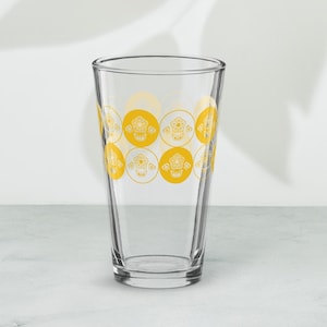 May include: A clear glass drinking glass with a yellow and white geometric pattern. The pattern features a repeating design of circles with a stylised flower inside each circle.