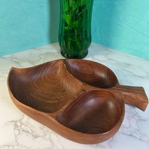 May include: A wooden leaf-shaped serving dish with three compartments. The dish is made of dark brown wood with visible grain patterns. A green glass vase with greenery is in the background.