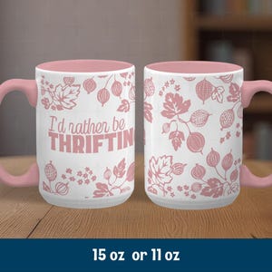 May include: White ceramic mug with a pink handle and interior. The mug features a pink floral design and the text "I'd rather be THRIFTIN" in pink. The mug is available in 15 oz or 11 oz sizes.
