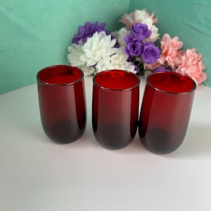 May include: Three stemless, ruby red drinking glasses. The glasses have a rounded shape and a dark base that fades to a lighter red at the rim. A bouquet of white, purple, and pink flowers is in the background.