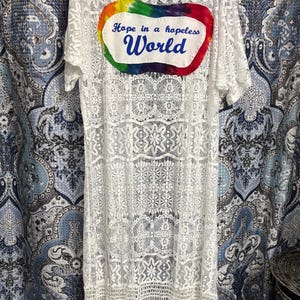May include: White lace kimono with a rainbow tie-dye patch that says "Hope in a hopeless World". The kimono has fringe at the bottom.