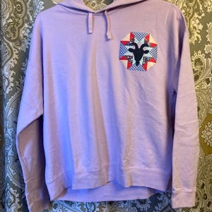 May include: A lavender hoodie with a patchwork design featuring a black goat silhouette on the front.