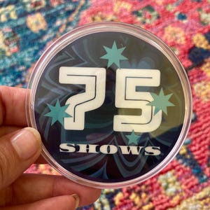 May include: A round, clear plastic button with a black and blue swirl design. The number 75 is printed in white with the word "SHOWS" below it. There are white stars with blue centers around the number.