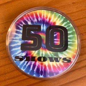 May include: A round, clear plastic button with a tie-dye pattern in shades of pink, blue, green, and yellow. The number "50" is printed in black on the button, along with the word "SHOWS".