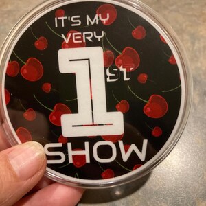 May include: A black and white round sticker with a white number one and the text "It's My Very 1st Show" surrounded by red cherries on a black background.