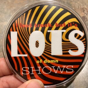 May include: A round, clear plastic button with a swirling orange, yellow, and black design. The text reads "I lost track, but it's LOTS of damn SHOWS" in white block letters.