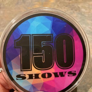 May include: A clear, round coaster with the number "150" and the word "SHOWS" printed on a colorful geometric background. The background features shades of blue, pink, and purple.
