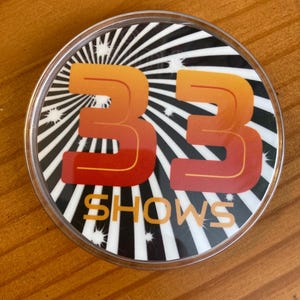 May include: A round, clear plastic button with a black and white striped background. The number 33 is in orange and red with a yellow outline. The words "SHOWS" are in yellow below the number.