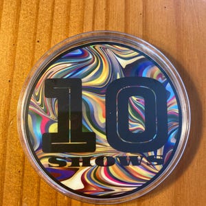 May include: A round, clear plastic coaster with a colorful, abstract swirl design. The number "10" is printed in black on the coaster, with the word "SHOWS" printed below it.