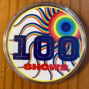 May include: A round, clear plastic button with a white background and a rainbow design. The number "100" is printed in blue and the word "SHOWS" is printed in red.