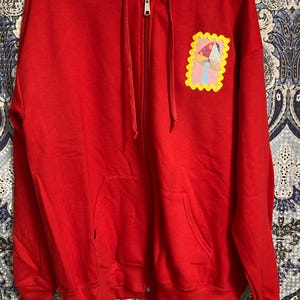 May include: A bright red zip-up hoodie with a hood and front pockets. A colorful patch featuring a mushroom design is on the left chest. The hoodie has a silver zipper and is made of a soft material.