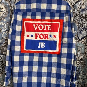 May include: A blue and white plaid flannel shirt with a red, white, and blue patch sewn on the back. The patch says "VOTE FOR JB" in white letters.