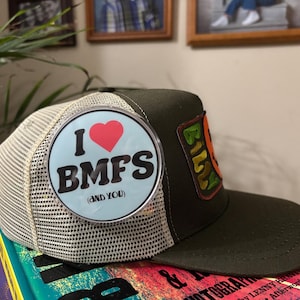 May include: A green and tan trucker hat with a patch that says "I ❤️ BMFS (and you)" in black text with a red heart. The hat also has a patch that says "Billy" in green and yellow.