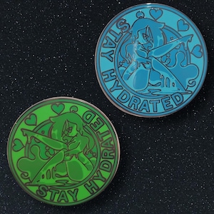 Op de afbeelding: Twee ronde emaille pins, één turquoise en één groen. Elke pin heeft een gestileerde illustratie van een vrouw met lang haar, met de woorden "STAY HYDRATED" rond de rand. De turquoise pin heeft een blauwe achtergrond en de groene pin heeft een groene achtergrond.