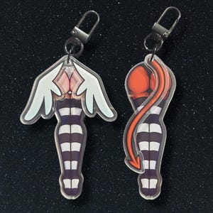 Puede incluir: Dos charms de acrílico con un personaje de dibujos animados con medias a rayas, uno con alas de ángel y el otro con una cola roja. Los charms están unidos a llaveros plateados.