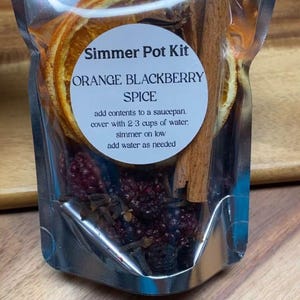 May include: A clear, resealable pouch containing a simmer pot kit with dried orange slices, a cinnamon stick, blackberries, and cloves. A white circular label reads: Simmer Pot Kit, ORANGE BLACKBERRY SPICE, add contents to a saucepan, cover with 2-3 cups of water, simmer on low, add water as needed.