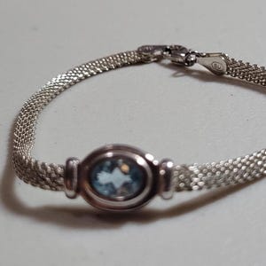 May include: A silver-toned mesh bracelet featuring a central oval-shaped light blue gemstone set in a smooth metal bezel. The bracelet includes a secure lobster claw clasp mechanism and a fine, flexible woven chain design that encircles the wrist.