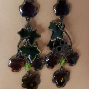 May include: A pair of dangle earrings with a floral design. The earrings are made of metal and have a dark brown, green, and purple enamel finish.