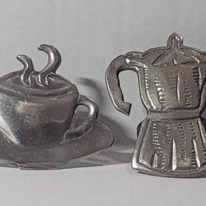 May include: Four silver-colored coffee-themed metal ornaments. Two are shaped like coffee cups with steam, and two are shaped like stovetop espresso makers. These decorative items are suitable for coffee lovers.