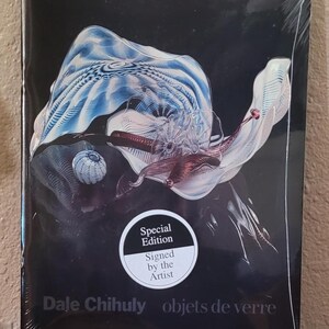 May include: A special edition book titled "objets de verre" by Dale Chihuly, featuring a colorful glass art design on a black background. The book is sealed in plastic and has a sticker that says "Signed by the Artist".