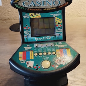 May include: A black and green 5-in-1 Deluxe Virtual Casino game with the text "Excalibur". The game features slots, blackjack, and poker. It has a digital display and various buttons for gameplay.