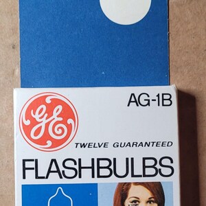 May include: Vintage GE AG-1B flashbulbs in a white and blue box. The box features the GE logo in red, the words "TWELVE GUARANTEED FLASHBULBS", and an illustration of a woman holding a camera. The blue cardboard backing has a circular sticker.