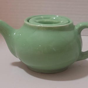 May include: A small, light green ceramic teapot with a matching lid. The teapot has a rounded body, a curved spout, and a handle. The lid has a small, circular handle. The teapot is approximately 4 inches long and 3 inches tall.