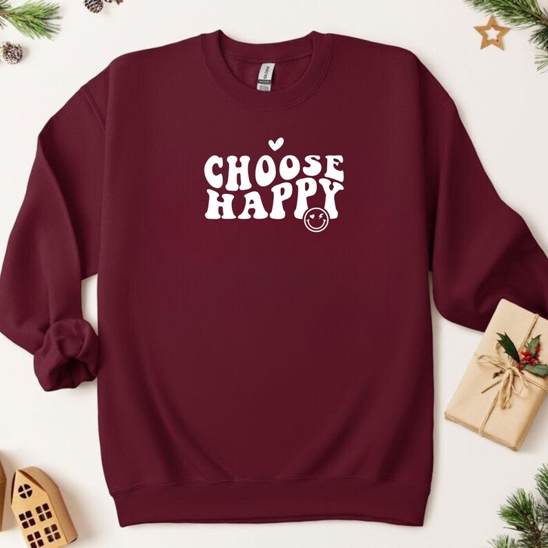 May include: Burgundy sweatshirt with white text that reads "Choose Happy" in a retro font. A small white heart and smiley face are above and below the text.
