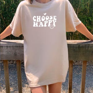 May include: A beige oversized t-shirt with the text "CHOOSE HAPPY" in a retro font with a white heart and smiley face.
