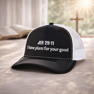 May include: Black and white trucker hat with the text "JER 29:11 I have plans for your good" in white. The hat has a black front panel and bill, with a white mesh back. The hat is sitting on a light-colored surface.