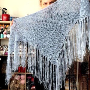 May include: A light blue triangular shawl crafted with a loose, open-knit texture. The garment features long, flowing fringe along the two bottom edges, creating a soft, airy appearance with a delicate, fuzzy yarn composition.