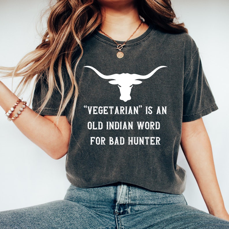 Puede incluir: Camiseta gris oscuro con un gr&aacute;fico de longhorn blanco y el texto "VEGETARIAN IS AN OLD INDIAN WORD FOR BAD HUNTER". La camiseta est&aacute; hecha de un material suave y c&oacute;modo.
