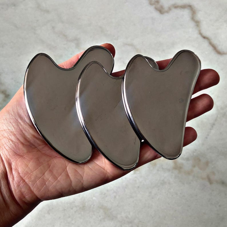 May include: Three silver-colored metal facial tools shaped like a heart. The tools are smooth and have rounded edges.