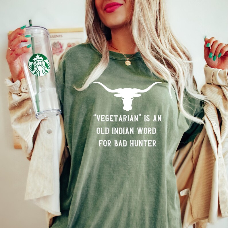Puede incluir: Camiseta verde oliva con un gr&aacute;fico de cuerno largo blanco y el texto "Vegetarian is an old Indian word for bad hunter". Un vaso transparente de Starbucks se sostiene en la mano izquierda.