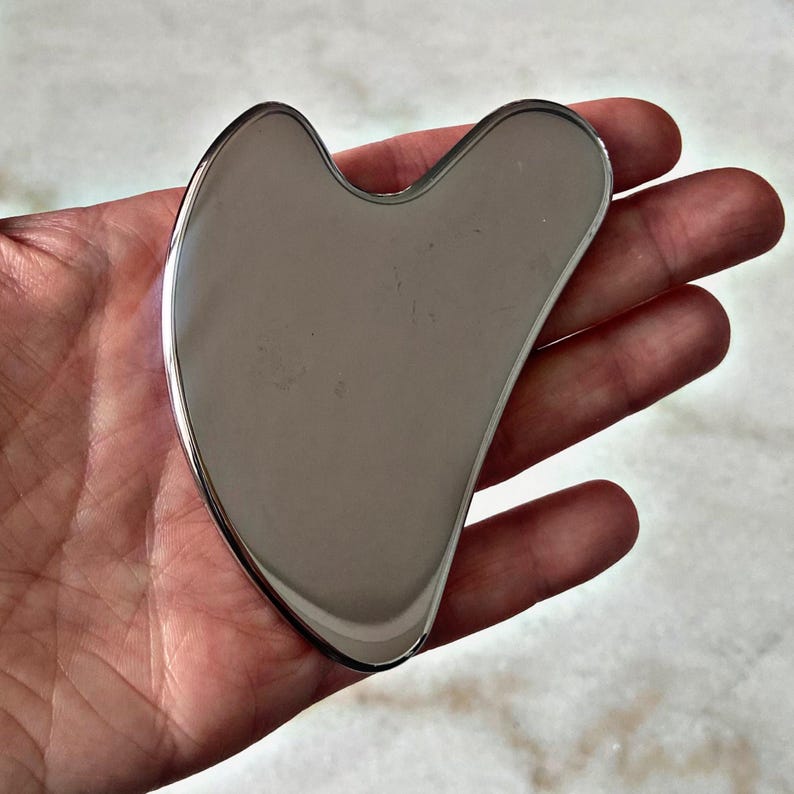May include: A silver-colored heart-shaped facial gua sha tool. The tool is smooth and has a rounded edge.