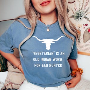 Puede incluir: Una camiseta azul con un gr&aacute;fico de longhorn blanco y el texto "Vegetarian is an old Indian word for bad hunter." La camiseta est&aacute; hecha de un material suave.
