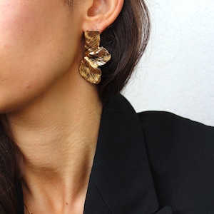 Puede incluir: Pendientes de color dorado en forma de flor con superficie texturizada. Los pendientes son usados por una persona con cabello oscuro y una chaqueta negra.