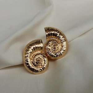 May include: A pair of gold-coloured earrings, each shaped like a spiral. The earrings have a textured surface and a shiny finish. They are displayed against a white background, which accentuates their design. These earrings are a fashion accessory.