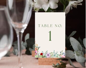 Pode incluir: Um cartão branco com texto verde que diz "TABLE NO. 1" com uma borda floral. O cartão está em um suporte de madeira.