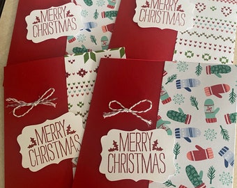 Puede incluir: Cuatro tarjetas de Navidad rojas con insertos de papel blanco. Los insertos presentan diferentes patrones, incluyendo guantes, copos de nieve y un diseño de punto de jersey. Cada tarjeta tiene una etiqueta blanca con el texto "Merry Christmas" en rojo.
