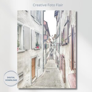 May include: A narrow, cobblestone street lined with white buildings with wooden doors and windows. The buildings have a faded, vintage look, and the street is empty.