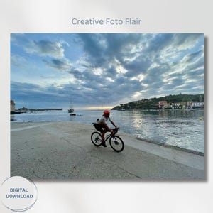 May include: A person rides a bicycle on a paved path along a waterfront with a town in the distance. The sky is cloudy and the water is calm.